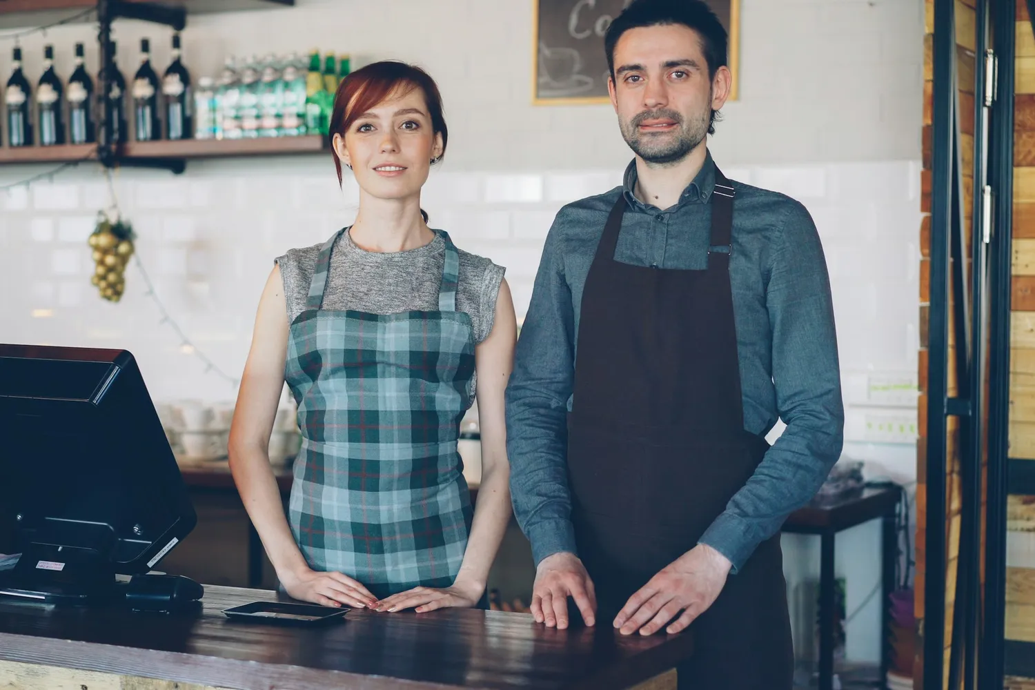Two café staff in aprons standing behind a counter beside a point-of-sale screen